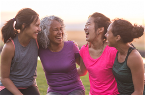 A group of four women of varying ages wearing brightly coloured active wear, huddled outdoors and laughing