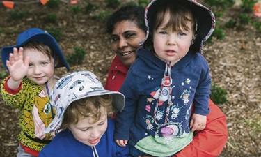 Three children smile and wave to the camera, one is being embraced by an educator