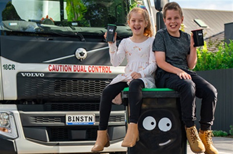 Two smiling children sitting on a green bin with cartoon eyes in front of a rubbish truck displaying a number plate with the letters BINSTN.