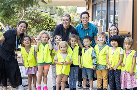 Four early learning educators standing outside a childcare centre with a group of children wearing high-vis vests