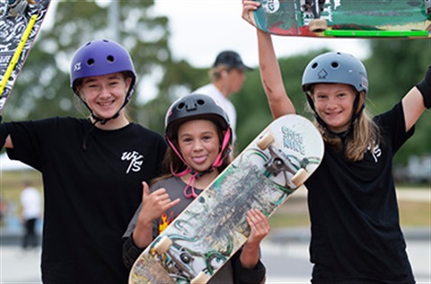 Three young people at a skate park, wearing helmets and holding up their skateboards