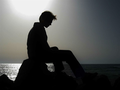 Man sitting with head bowed near the ocean.