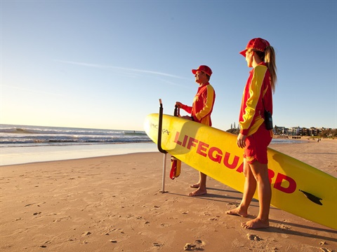 Two lifeguards stnading on the beach looking out at the water. One of them is holding a yellow paddleboard which reads 'Lifeguard' in red lettering